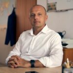 A confident adult man in a white shirt sitting in a well-lit office with a thoughtful expression.